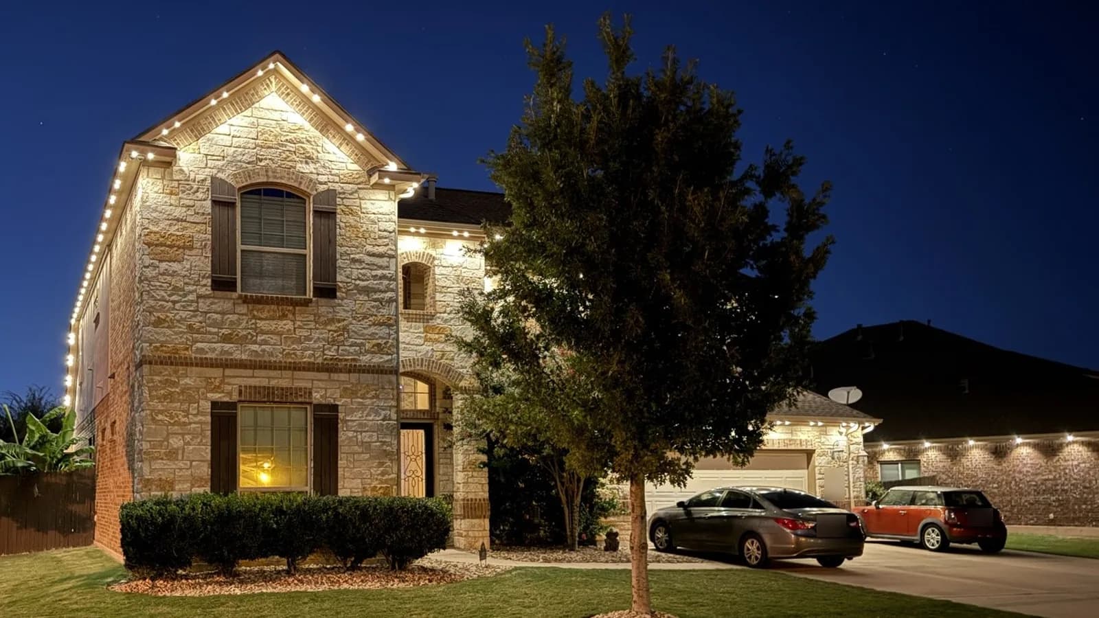 Two neighboring homes at night: an even warm white glow on the stone home at left and separate bright points with shadows between them on the brick home at right