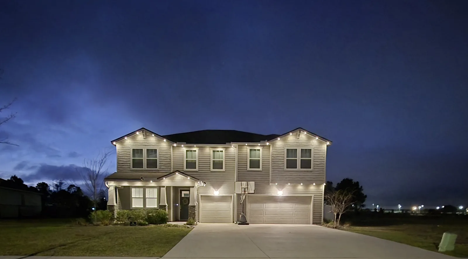 Two-story Green Cove Springs home at night with warm white permanent lighting along the upper and lower rooflines and a three-car garage.